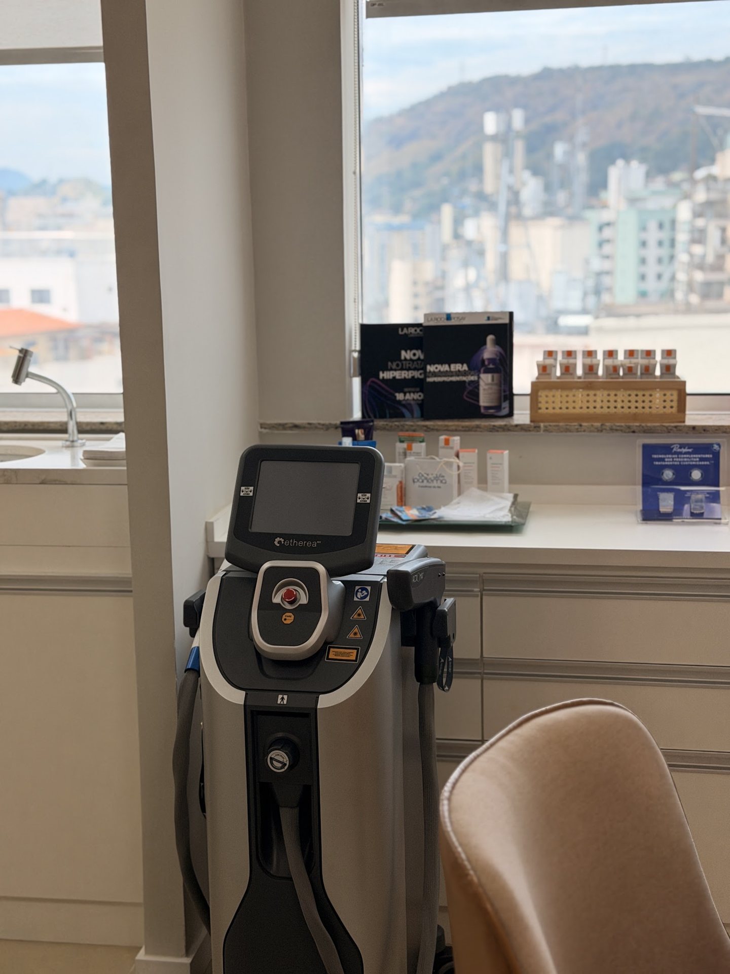 Sala de procedimento do consultório, com aparelho de coluna, suporte dourado com ampolas e a janela com vista do morro de Niterói